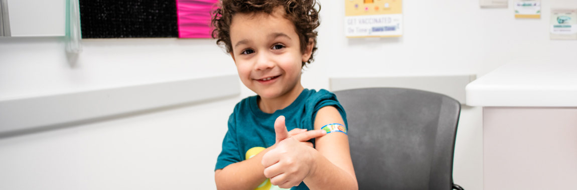 A child giving a thumbs up after getting a vaccine. He has a band-aid on his arm.