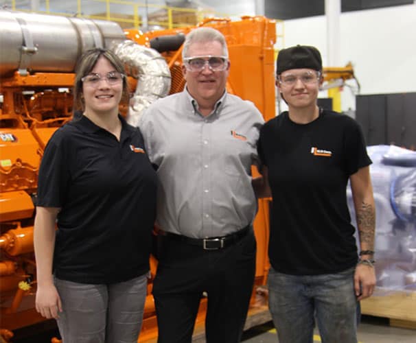 A photo shows two young women, past participants of Oakland County Manufacturing Day, with an older man in the middle.