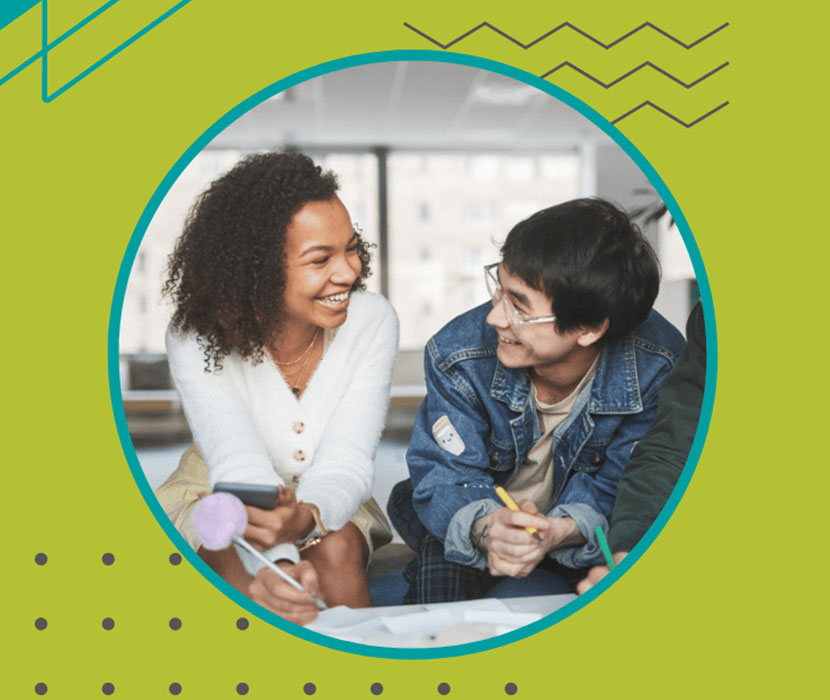 A young black woman student and young Asian student man sit on a couch, sharing smiles and laughter, enjoying a fun moment together holding pens in their hands.