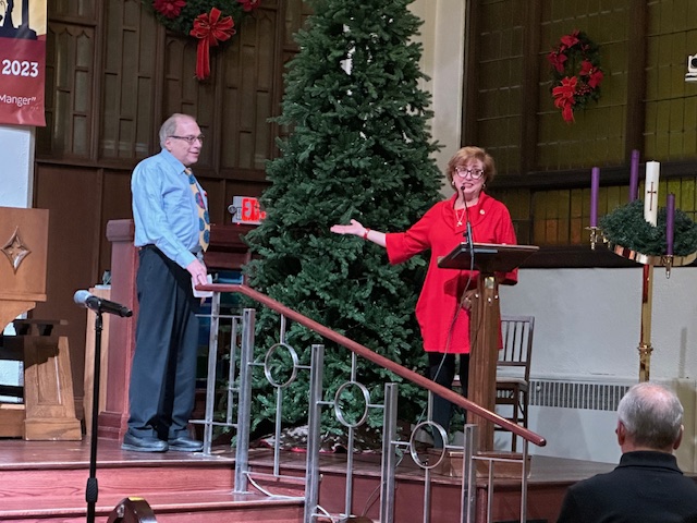 A person speaking at a church. The person is directing people to look at another individual on stage. There is a Christmas tree in the background.