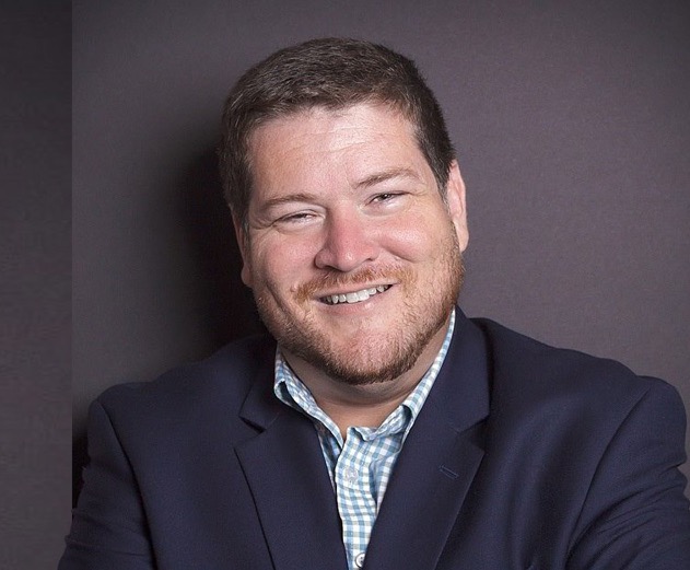 An image shows a professional headshot of a man, John Bry. He is wearing a business suit and a plaid collared shirt. 